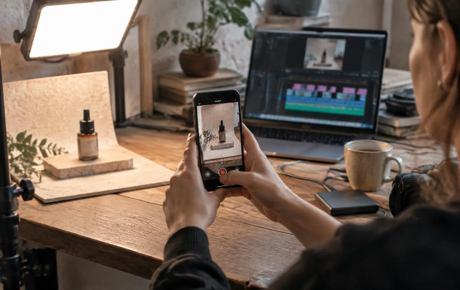 Designer filming a product on a phone in a small tabletop set, with a video edit timeline open on a laptop